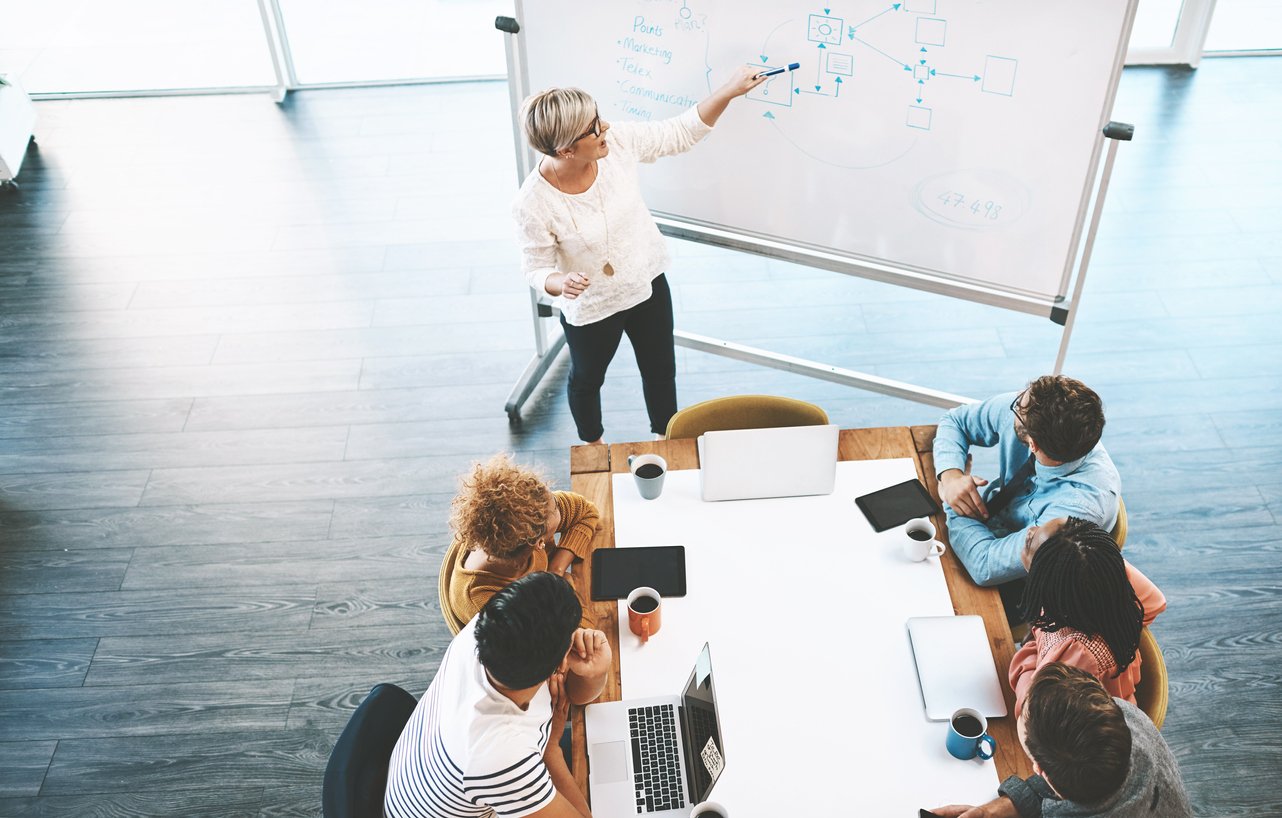 business-meeting-with-people-sitting-around-table-and-woman-speaking-while-standing-at-white-board