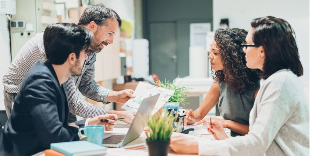 four-coworkers-sitting-around-table-in-office-with-laptop-papers-and-small-potted-plant